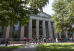 Students walk on a pathway through green lawns. In the back ground is a majestic library of red brick with off-white pillars