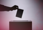 A silhouetted hand places a ballot into a ballot box.