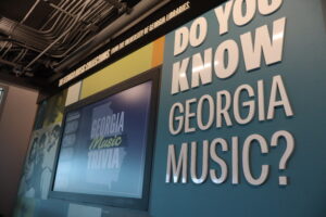 A closeup of a blue sign with white text that says "Do you know Georgia music?" There is a computer screen advertising trivia to the left.